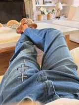 Pantalón de jean azul con corte holgado y detalle de tres cruces bordadas en el muslo derecho. Presenta costuras reforzadas y cierre frontal con botón metálico.