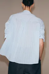 Camisa de corte oversize con estampado de rayas verticales, cuello redondo tipo mao y cierre frontal con botones. Presenta manga larga con puños anchos, un bolsillo de plastrón con solapa en el frente y bajo asimétrico.