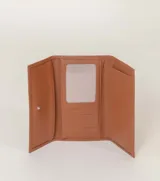 Billetera tipo sobre color champagne, con textura de cuero granulado y cierre de solapa. Presenta una placa metálica rectangular con el nombre de la marca grabado en el frente.