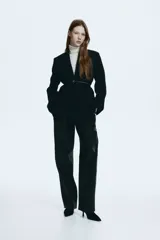 Blazer de sarga color negro con solapas de muesca, frente cruzado y cinturón fino ajustable con hebilla metálica. Presenta bolsillos delanteros ribeteados con solapa y diseño forrado.