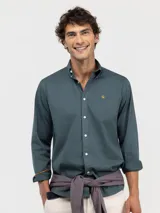 Camisa de hombre color azul, con cuello buttondown, cierre frontal con botones y logo bordado en el pecho. Presenta un acabado vintage logrado mediante el proceso de teñido en prenda.
