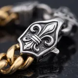 Pulsera de eslabones gruesos con baño de oro, destacada por un cierre central de diseño ornamental en forma de flor de lis con acabado plateado.