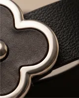 Cinto de cuero negro con hebilla plateada en forma de trébol de cuatro hojas, decorada con esmalte negro brillante y un detalle metálico esférico en el centro.