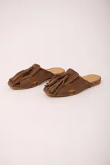 Zueco de cuero color marrón, estilo mule, con puntera cuadrada y tira ancha adornada con flecos. Suela plana.