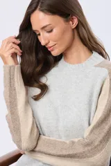 Buzo tejido de abrigo con cuello redondo y manga larga. Presenta un diseño de bloques de color en tonos gris claro y beige.