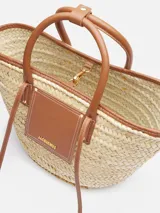 Bolso tipo canasto tejido a mano en fibra vegetal con detalles de cuero marrón, asas cortas enrolladas y logotipo Jacquemus en letras doradas en el frente.