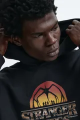 Canguro negro con capucha y bolsillo frontal tipo canguro. Presenta un estampado gráfico en el pecho con el logo de la serie Stranger Things y una silueta del Mind Flayer sobre un atardecer.