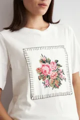 Remera color crudo de manga corta, con cuello redondo y un aplique bordado frontal con diseño floral enmarcado por pespuntes.