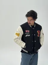Campera estilo varsity con cuerpo negro y mangas blancas. Presenta parches bordados en el pecho y las mangas, cierre frontal con botones a presión y puños, cuello y ruedo acanalados con rayas blancas y negras.