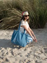 Bolso tipo tote confeccionado en tela denim de color azul claro. Presenta un diseño amplio con costuras visibles, doble asa de hombro y un detalle bordado circular en color rojo en la parte inferior.