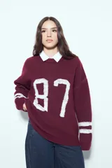 Sweater de punto color bordó con diseño de estilo universitario, presenta el número 87 estampado en blanco en el frente y rayas horizontales blancas en las mangas. Posee cuello redondo y corte holgado.