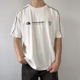 Remera oversize blanca de manga corta con detalles de líneas negras en las costuras de los hombros y mangas. Presenta un gran estampado en la espalda con el número 99 en diseño rayado y el logo de la marca en la parte superior.