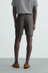 Bermuda de hombre color beige claro, corte regular fit, con pliegues frontales, bolsillos delanteros y bolsillos traseros de doble vivo con botón.