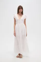 Vestido largo blanco de algodón con escote en V, manga corta con volados de broderie y talle entallado con botones frontales. La falda es amplia y presenta detalles de bordados calados en el ruedo.