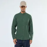 Camisa de manga larga de franela verde oscuro, con cuello mao y cierre frontal de botones marrones. Posee un bolsillo de parche en el pecho izquierdo.