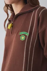 Buzo de tejido de punto color marrón con cuello alto y cierre frontal parcial. Presenta rayas verticales finas en color claro, puños y ruedo en tejido acanalado color crema, y detalles en color verde lima en el tirador del cierre, el cordón de ajuste del ruedo y el parche bordado en el pecho.