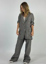 Blazer de lino color beige con finas rayas verticales oscuras. Presenta solapas de muesca, dos botones frontales y bolsillos de parche en la parte inferior. El borde de las solapas y los puños tiene un ribete oscuro contrastante.