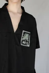 Remera oversize negra de piqué de algodón con bolsillo estampado con una palmera Phoenix Canariensis. Tiene elástico verde con hebilla de ajuste en la parte inferior.