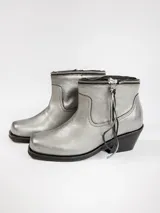 Bota de cuero metalizado plateado con punta cuadrada y tacón bajo. Presenta un diseño versátil con cierre metálico que permite desmontar la polaina superior, transformando la silueta de bota alta a botín.