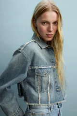 Chaqueta de jean estilo crop, con cuello solapa, manga larga y cierre frontal con botones metálicos. Presenta bolsillos de plastrón con solapa en el pecho y detalles de costuras deshilachadas en el frente.