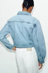 Cazadora corta de denim color celeste con cuello solapa, manga larga y hombreras. Cuenta con bolsillos delanteros con solapa y cierre frontal de botones metálicos.