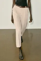 Pantalón tobillero de tiro medio, color beige, con pinzas delanteras y bolsillos frontales. Incluye un cinturón fino de cuero marrón con hebilla metálica dorada y cierre frontal con cremallera, botón interior y gancho.