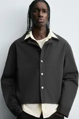 Sobrecamisa negra de algodón con estructura canvas, corte cropped y calce holgado. Presenta cuello solapa, manga larga, bolsillos laterales en cadera y cierre frontal con botones a presión metálicos.