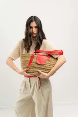 Bolso tipo canasto de paja con detalles en cuero rojo, incluyendo un bolsillo externo con solapa y cierre con botón a presión, y correa regulable.