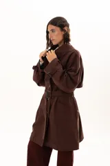 Trench color chocolate con cuello alto, cierre frontal con botones y cinturón ajustable en la cintura. Presenta mangas largas con puños ajustables mediante tiras y hebillas, y bolsillos frontales.