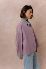 Camisa lila de calce relajado y manga larga, con cuello clásico, puños y bolsillo delantero en denim contrastante. Cierre frontal con broches metálicos.