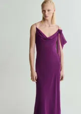 Vestido largo de fiesta color violeta, con diseño recto, cuello drapeado y tirantes finos. Presenta un detalle de flor con drapeado en uno de los hombros y cierre de cremallera lateral.