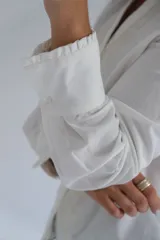 Blusa blanca de manga larga con cuello mao y cierre frontal de botones. Presenta detalles de bordados decorativos en los laterales del frente y puños anchos.