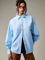Camisa de rayas verticales celestes y blancas, con cuello clásico, mangas largas abullonadas con puños abotonados y espalda abierta con lazo regulable.