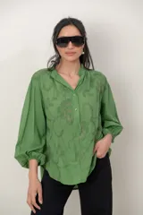 Camisa de algodón verde con bordados al tono, escote en V y cuello camisero corto, mangas tres cuartos abullonadas con puños elásticos y cierre frontal parcial con botones.