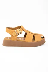 Sandalia tipo guillermina de cuero color camel, con diseño de tiras anchas entrelazadas, cierre con hebilla metálica lateral y suela de plataforma plana en tono marrón.