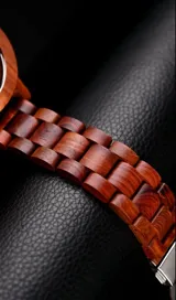 Reloj de pulsera con caja, esfera y malla de madera de sándalo rojo, con marcadores y manecillas negras. Presenta un diseño circular con corona lateral.