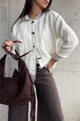 Cardigan de punto trenzado color natural, con cuello redondo, cierre frontal de botones negros y mangas largas con puños acanalados.