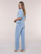 Camperita de denim color celeste con efecto lavado suave, cuello camisero clásico y cierre frontal de botones metálicos. Tiene mangas cortas con fruncido en los hombros, bolsillos delanteros bajos y costuras estructuradas.