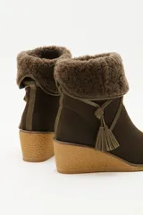 Bota de caña media color verde oliva, confeccionada en cuero con detalle de solapa superior en lana rizada. Presenta un cordón decorativo con borlas en el mismo tono y una suela de plataforma tipo cuña en color beige.