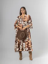 Vestido largo de manga tres cuartos, con cuello camisero y botones frontales. Presenta un estampado abstracto en tonos marrón oscuro, beige y blanco. El calce es amplio y tiene un ruedo con volado.