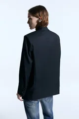 Blazer de corte oversize confeccionado en sarga, con hombreras marcadas, solapas clásicas y cierre frontal con un botón forrado a tono. Presenta dos bolsillos delanteros con solapa, logo bordado en una de las tapas y bordado decorativo en el cuello trasero. El diseño cuenta con un largo irregular que llega por debajo de la cadera y está completamente forrado.