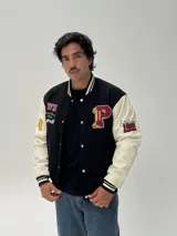 Campera estilo varsity con cuerpo negro y mangas blancas. Presenta parches bordados en el pecho y las mangas, cierre frontal con botones a presión y puños, cuello y ruedo acanalados con rayas blancas y negras.