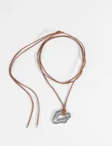 Collar largo con cordón de gamuza marrón y dije de acero quirúrgico con forma irregular y diseño de líneas concéntricas.