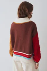 Cardigan de punto multicolor con diseño colorblock, cuello polo rosado con borde rojo, cierre frontal de botones, mangas largas abuchonadas (una roja y otra verde militar con tejido de ochos) y detalles de rayas en los puños y el ruedo.