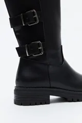 Bota de caña alta tipo biker en símil cuero color marrón oscuro. Presenta múltiples tiras horizontales con hebillas metálicas laterales y una suela robusta con relieve.