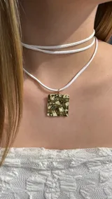 Gargantilla tipo choker compuesta por un cordón de gamuza blanca de doble vuelta y un dije cuadrado de metal dorado con textura irregular y efecto martillado.