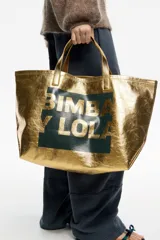 Bolso tipo tote de Bimba y Lola, color dorado con acabado metalizado y efecto arrugado. Presenta un diseño cuadrado con estampado central de la marca en un recuadro oscuro y doble asa de mano en color crema.