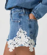 Short de jean tiro medio con encaje floral blanco aplastado en los laterales y dobladillo deshilachado. Cuenta con bolsillos delanteros y traseros, pasacintos y cierre frontal con botón y cremallera.
