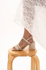 Sandalia de cuero con acabado metalizado dorado, diseño de doble tira delantera y cierre con hebilla ajustable en el tobillo. Cuenta con taco cuadrado y puntera cuadrada.