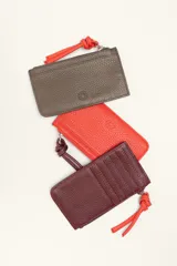 Monedero rectangular de cuero vacuno con textura granulada en color marrón topo. Cuenta con cierre superior metálico y un tirador decorativo de cuero en color naranja con un nudo. Presenta un pequeño logo grabado en el centro.